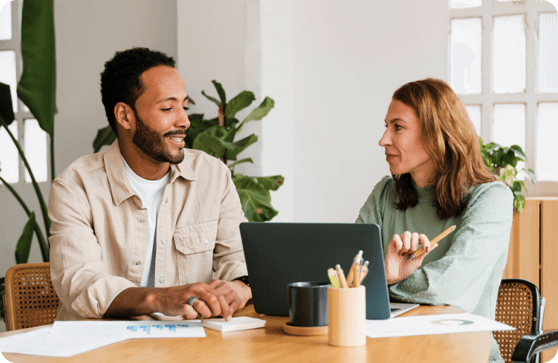 Two people sit next to each other in front of a table, on which there are a laptop, some pens and some pieces of paper