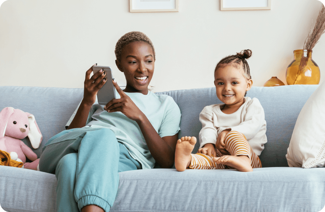 A woman who is holding a smartphone and a child sit next to each other on a couch while smiling