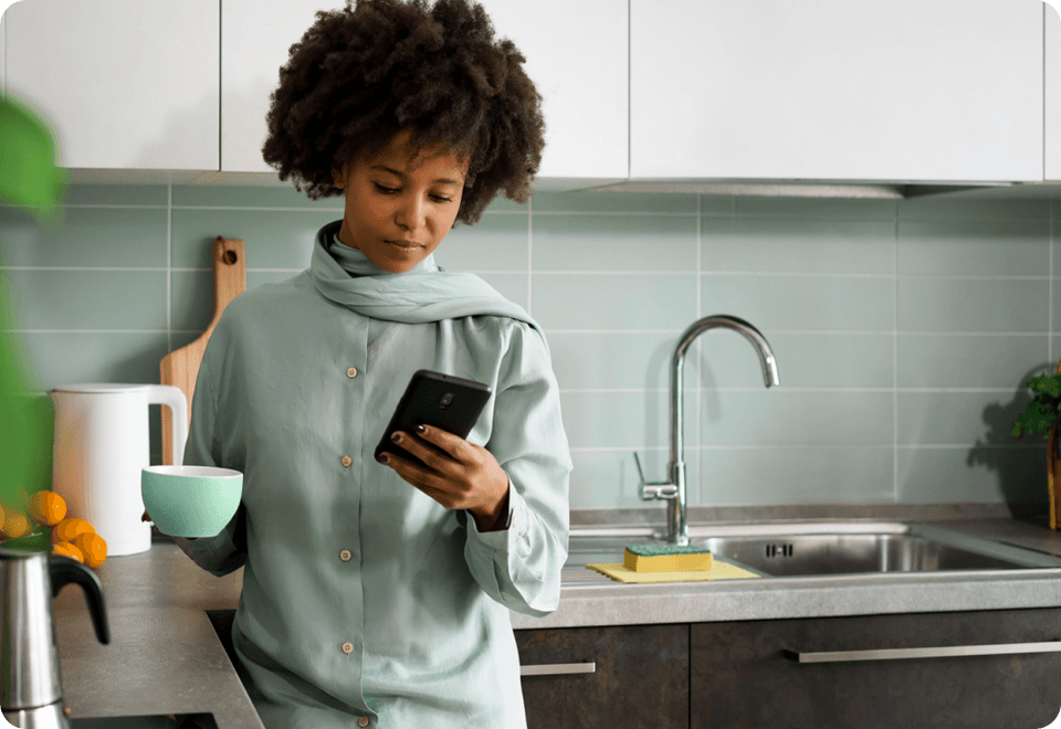 A woman in front of a kitchen sink holds a mug while looking at a smartphone