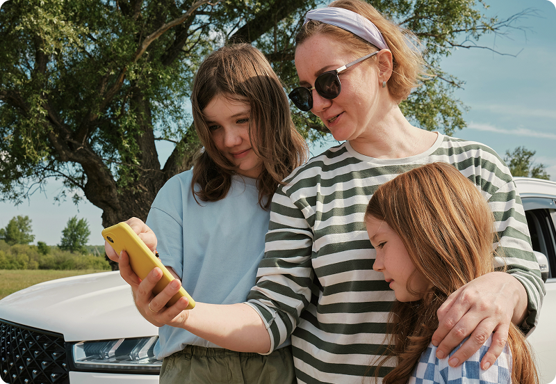 Mother with two daughters standing next to a broken car, looking at a phone, smiling.