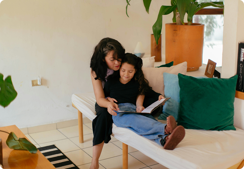 Mother and daughter sitting on a couch reading a book together.