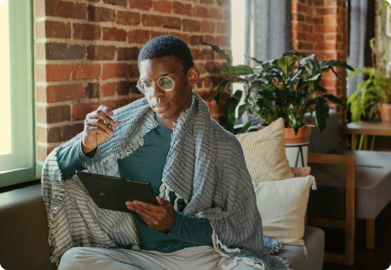 Person sitting comfortably on a couch at home, wrapped in a blanket, looking at a tablet.