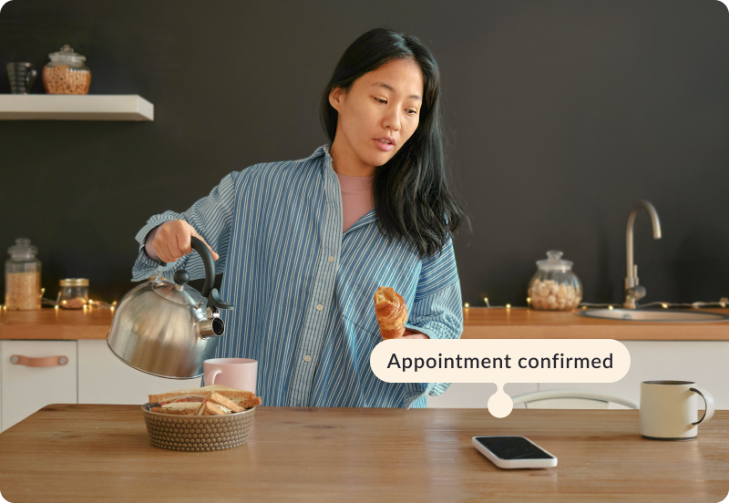 Person making a morning coffee in a kitchen, looking on phone. The tag next to the phone reads "Appointment confirmed"