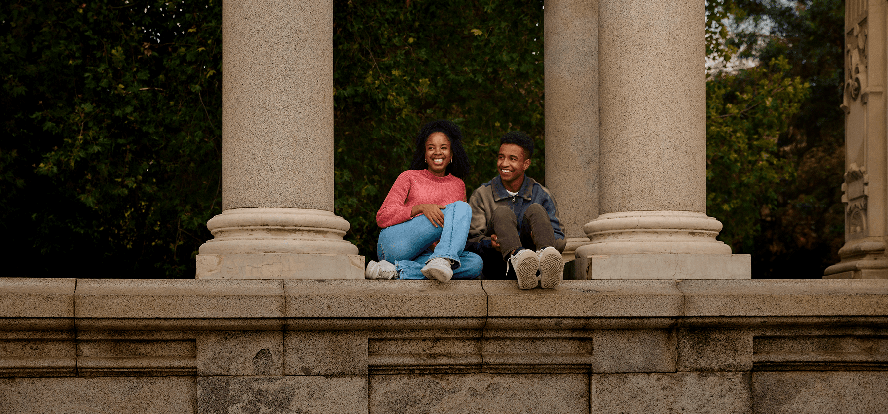 Two young people sitting and laughing together between grand columns surrounded by autumn trees in Ireland.