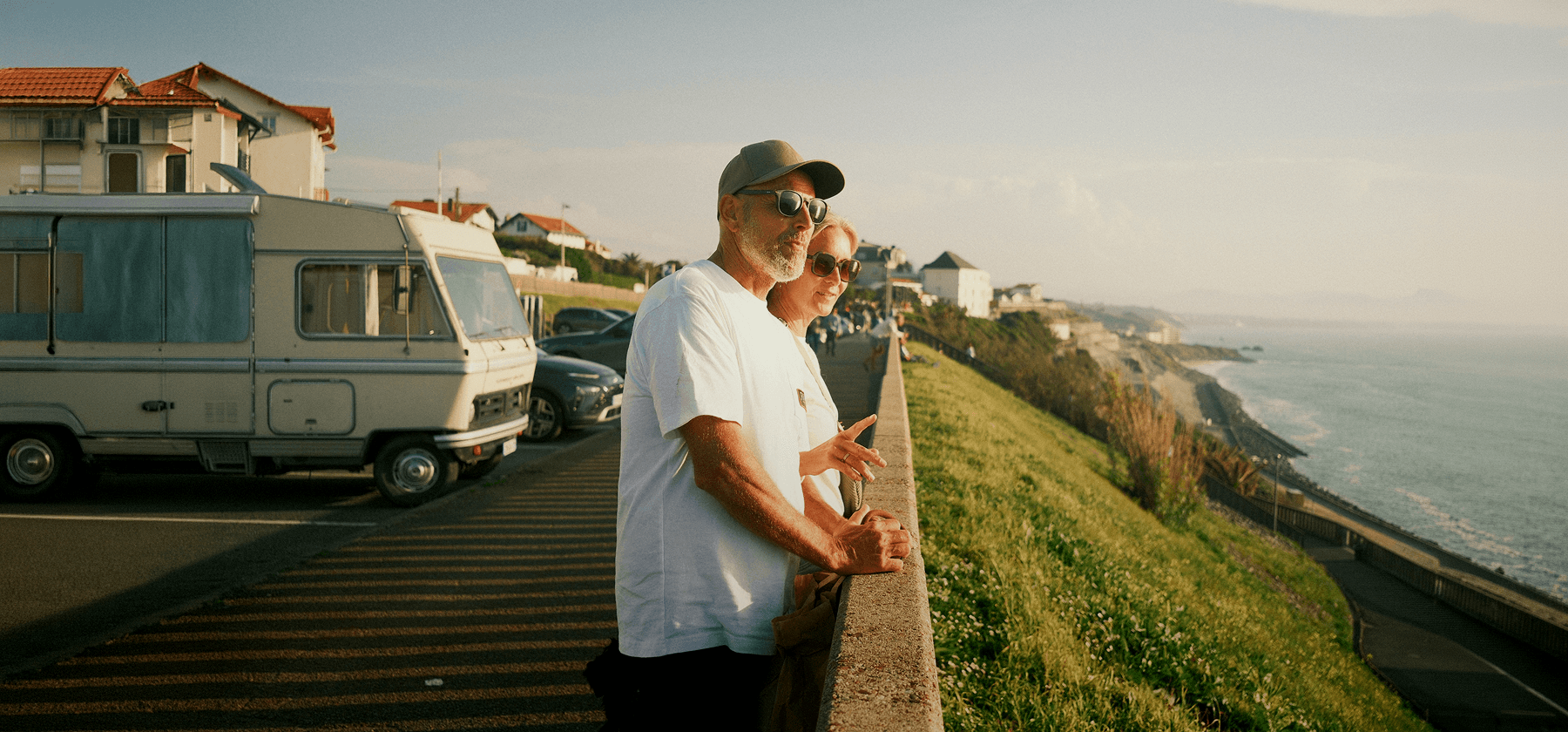 An older couple wearing sunglasses leaning on a wall overlooking a coastal road and the sea at sunset in France.