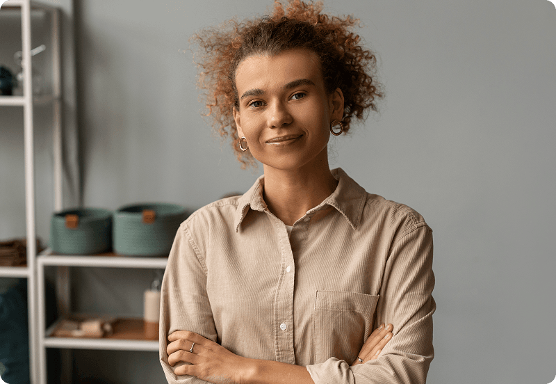 Image of a woman with curly hair, smiling confidently with arms crossed, wearing a beige shirt in a minimalist room.