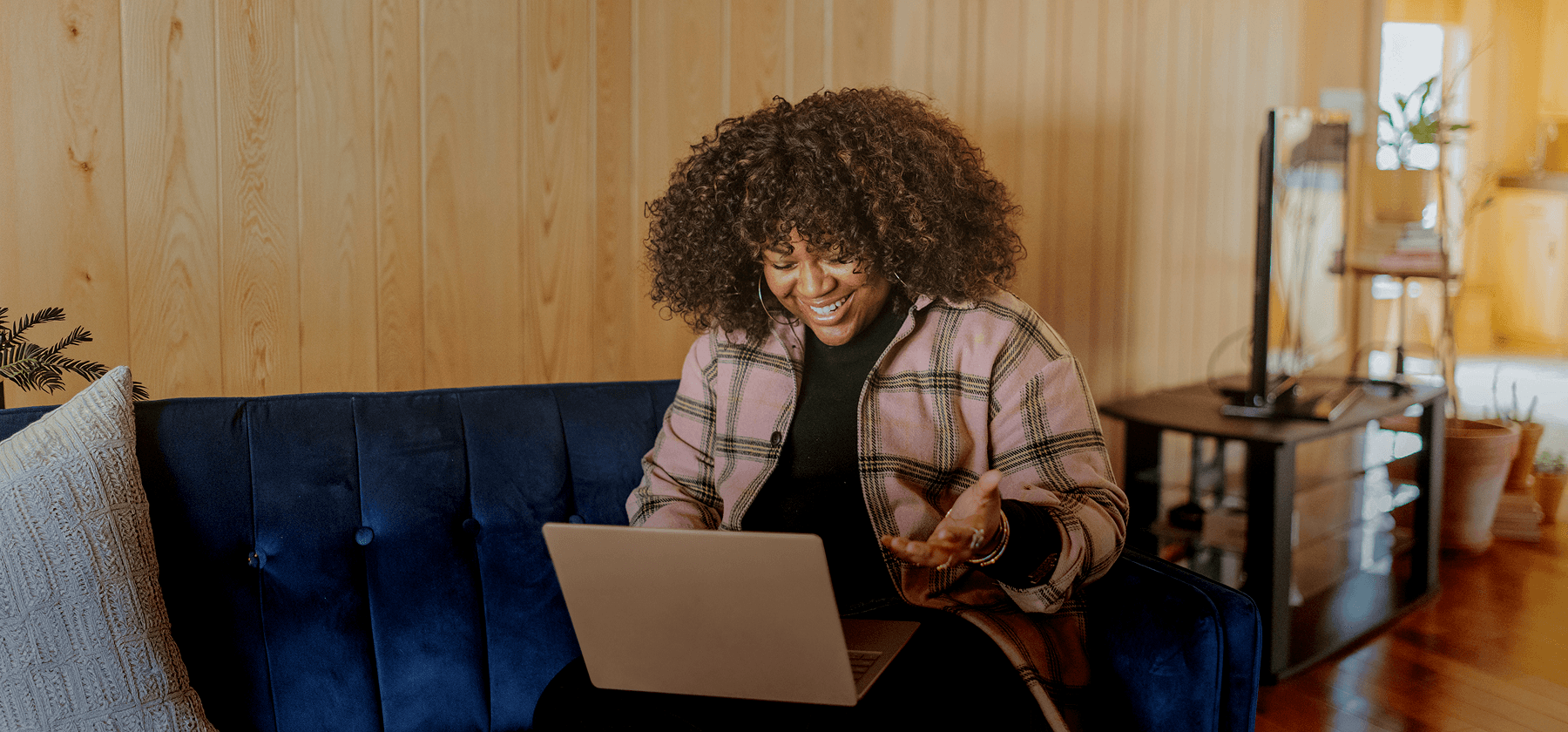 A woman with curly hair wearing a plaid jacket sitting on a blue velvet sofa, smiling and gesturing while on a video call on her laptop in a warm wooden-panelled living room.