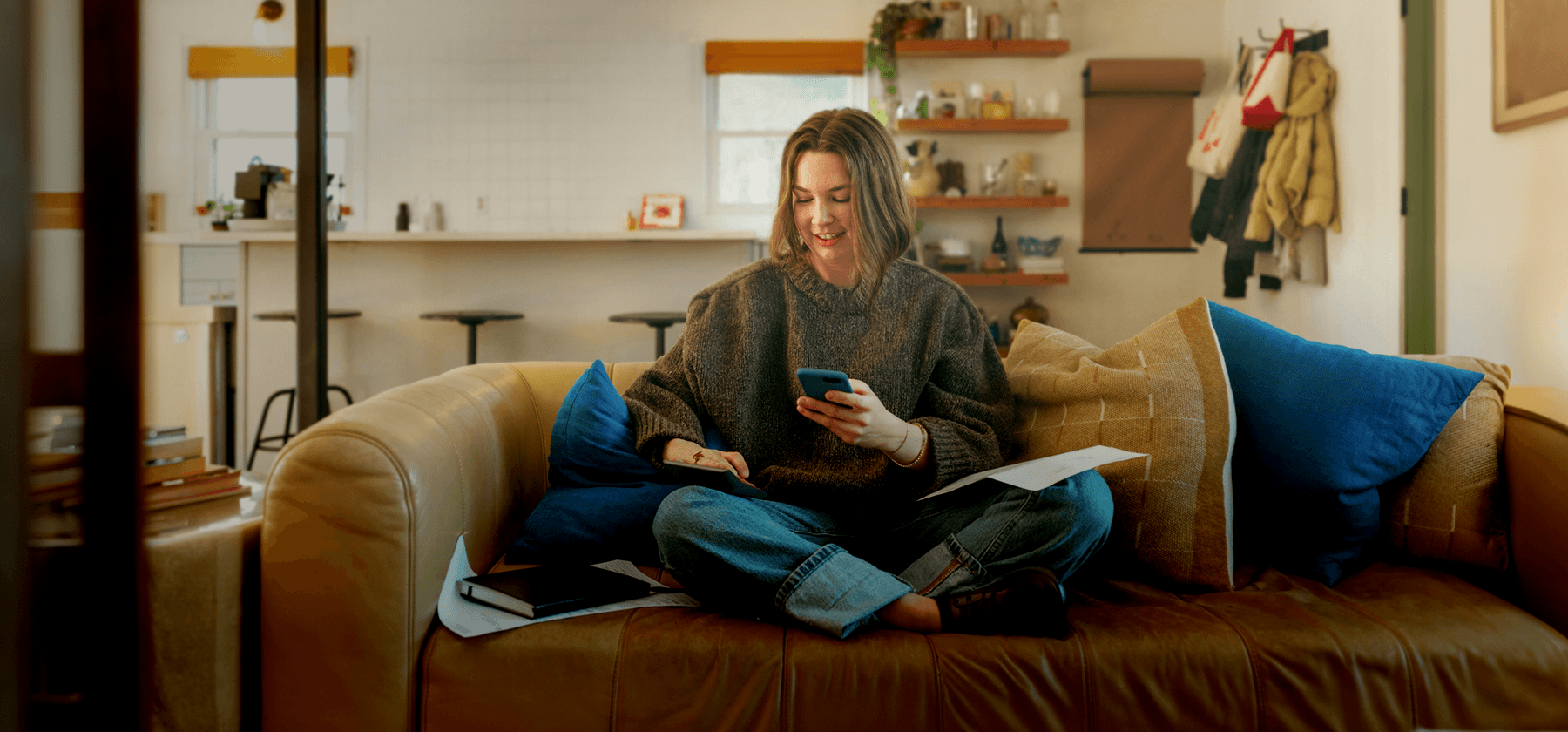 A woman sitting cross-legged on a sofa, smiling as she looks at her phone with papers in her lap.