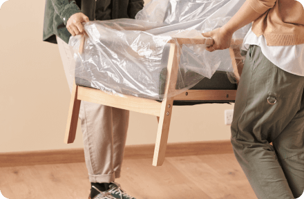Two people carrying a partially wrapped wooden sofa inside a room with wooden flooring