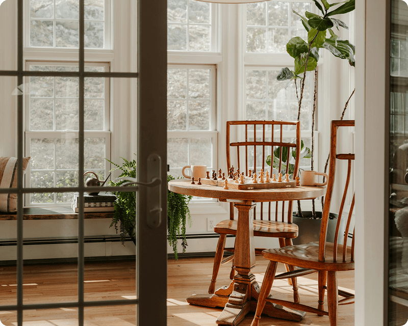 A cosy sunlit room with a wooden table set up for a chess game, surrounded by plants and large windows overlooking greenery in Germany.