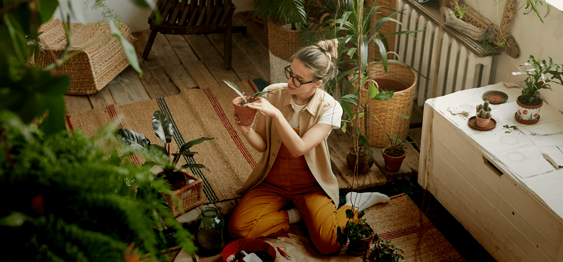 A woman wearing glasses sitting cross-legged on the floor of a plant-filled home, carefully tending to a potted plant surrounded by lush indoor greenery in Germany.