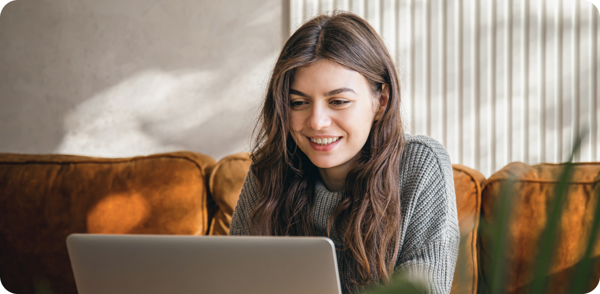 Young woman sitting on a couch using a laptop, smiling, with soft natural light filtering into the room.