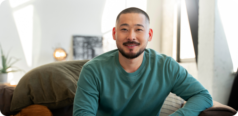 Man with a beard and green sweatshirt sitting on a couch in a sunlit living room, smiling at the camera.
