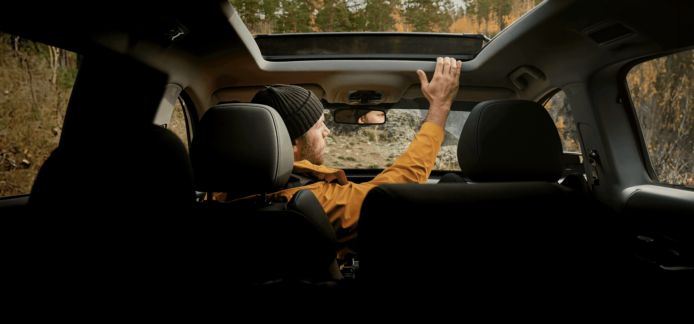 A man in a yellow jacket and beanie hat sitting in the driver's seat of a car, looking out the window at a forest landscape in France.