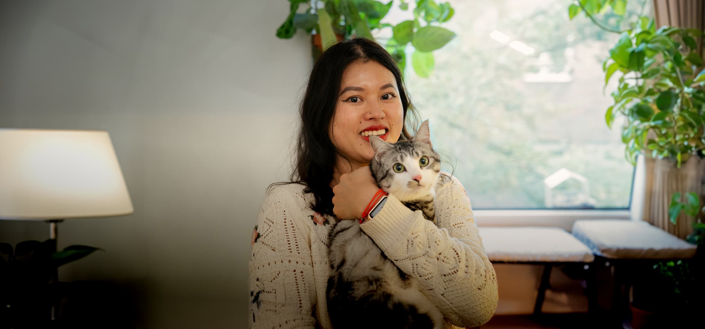 A woman smiling warmly while holding her tabby cat close in a cosy, plant-filled living room.