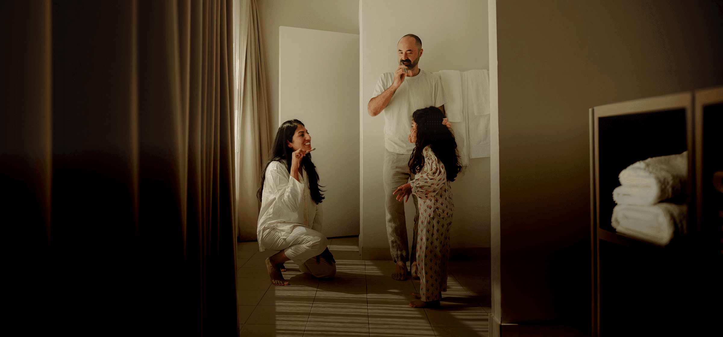 A family brushing their teeth together in a warmly lit bathroom — a mother crouching down to her child's level while a father stands nearby.