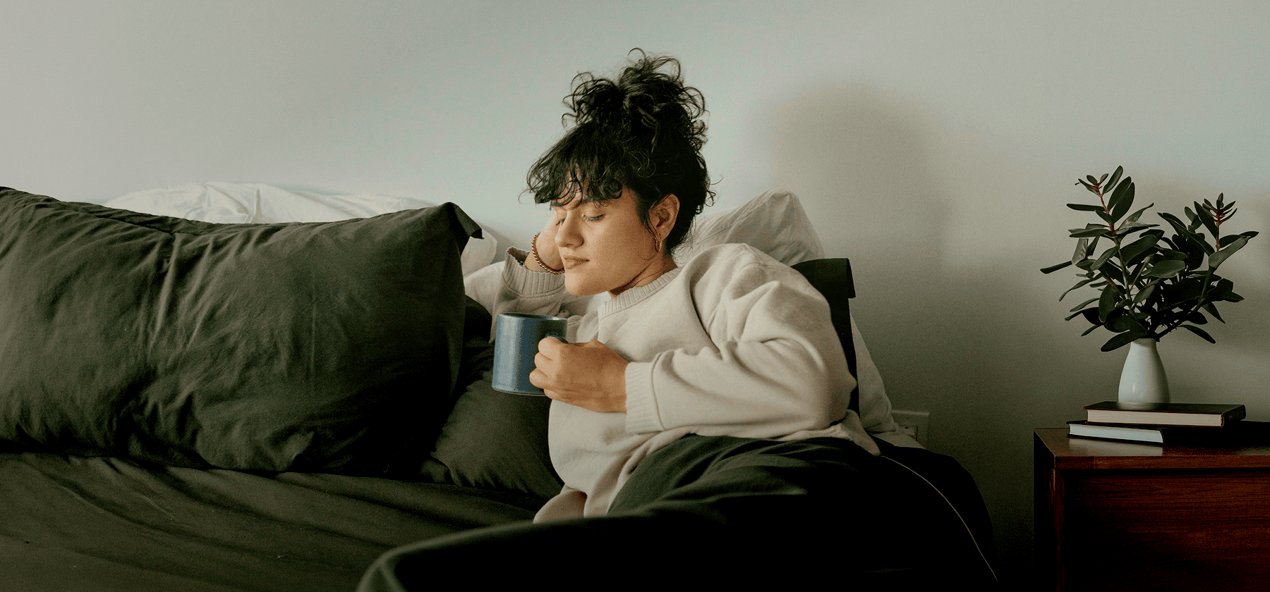 A woman with curly hair sitting in her bed, holding a mug and resting in a calm bedroom in Germany.