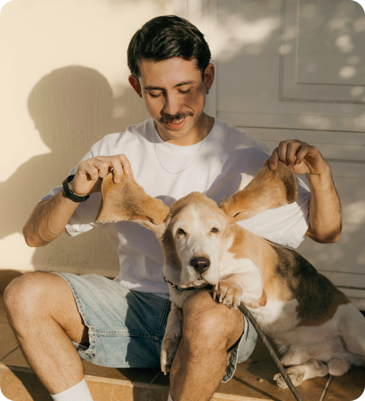 Persona sentada afuera con su perro, sonriendo y jugando con las orejas del perro.