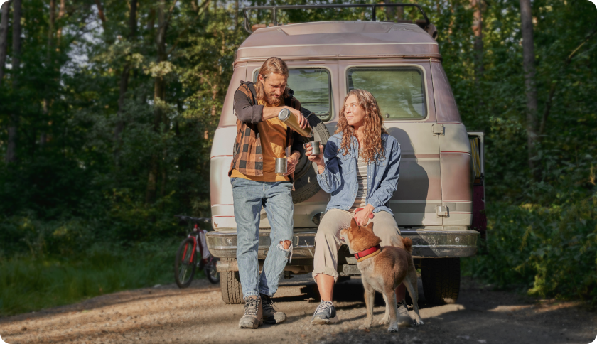 Una pareja con su perro sentada frente a su furgoneta en un bosque, tomando café de un termo.