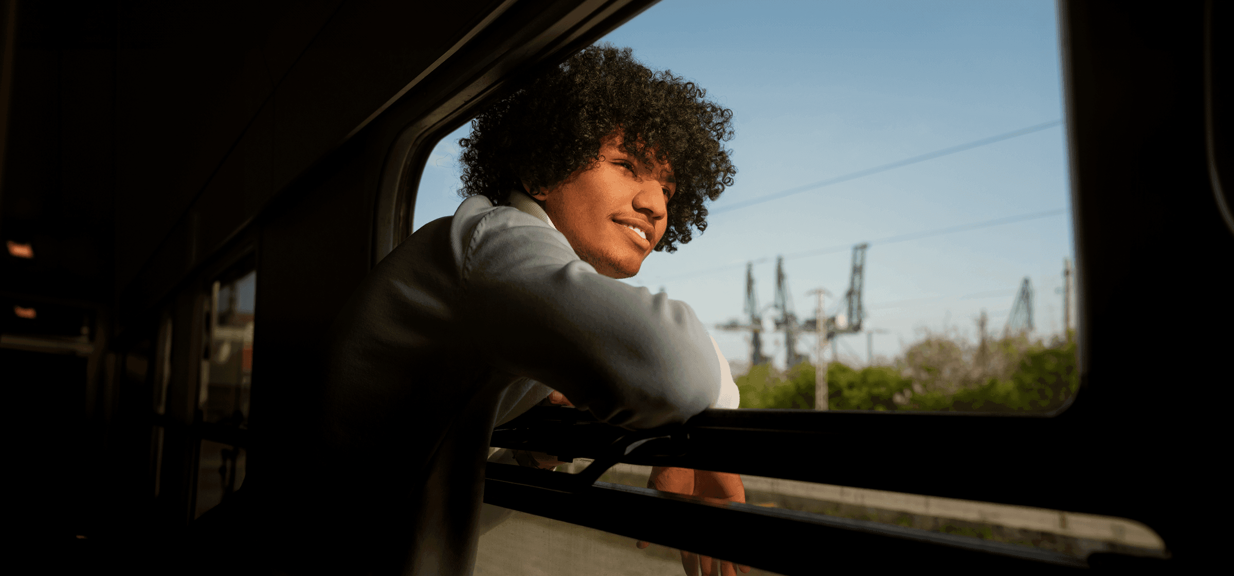 A young man leaning out of a train window, smiling as he looks into the distance on a sunny day.