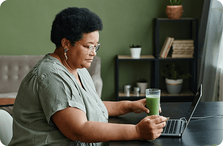 Persona jubilada sentada frente a su computadora portátil en casa y bebiendo un batido.