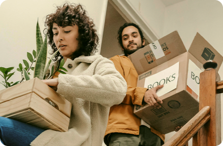 A couple holding moving boxes with books and plants, walking down the stairs and moving out from their old apartment.