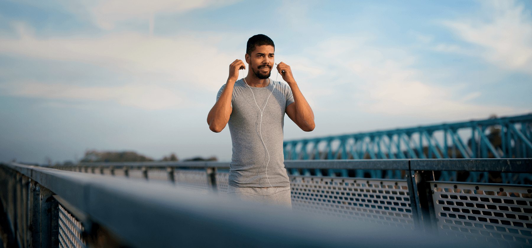 A man in a grey t-shirt putting in earphones while standing on a bridge outdoors on a sunny day in Portugal.