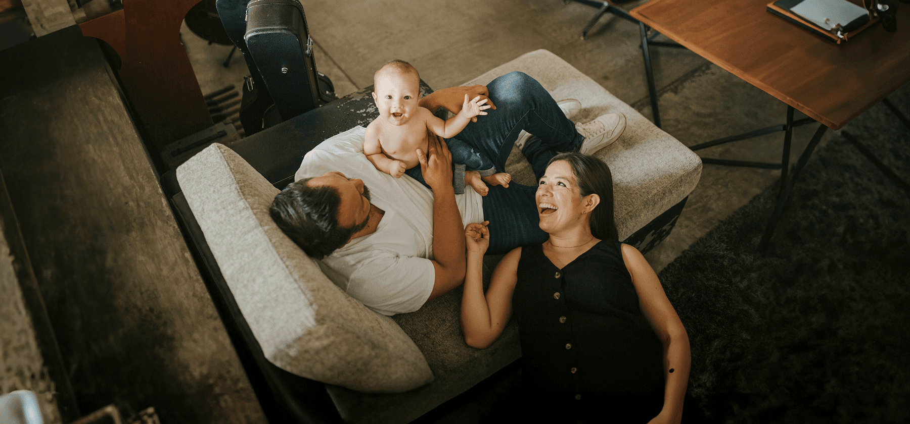A happy young family with a laughing baby lying on a parent's chest on a sofa, viewed from above, in France