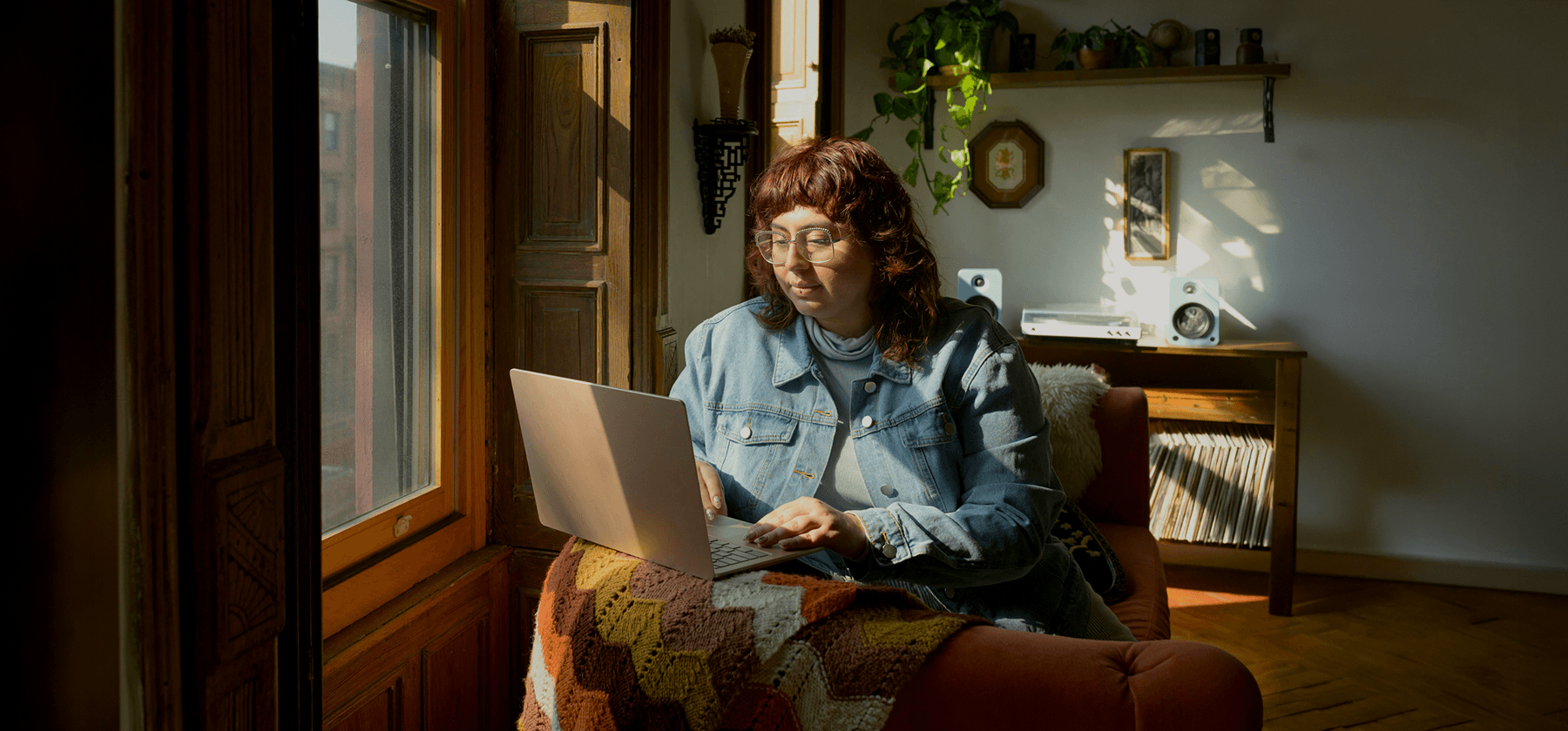 A woman with curly red hair working on a laptop by a sunny window in a cosy home office in France