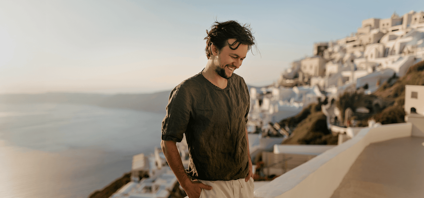 A smiling young man standing on a terrace with the iconic white-washed buildings and sea views of Santorini in the background in Greece.
