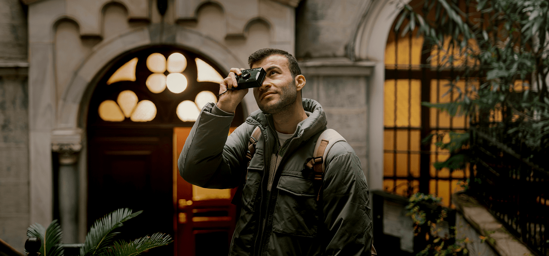 A young man with a backpack holding a film camera up to his eye while exploring an ornate historic building in Italy.