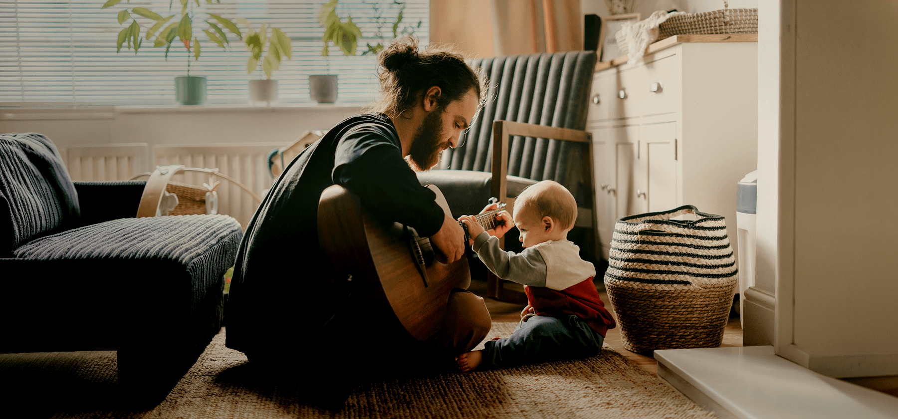 A bearded father sitting on the floor playing guitar for his curious baby in a cosy sunlit living room in Portugal.