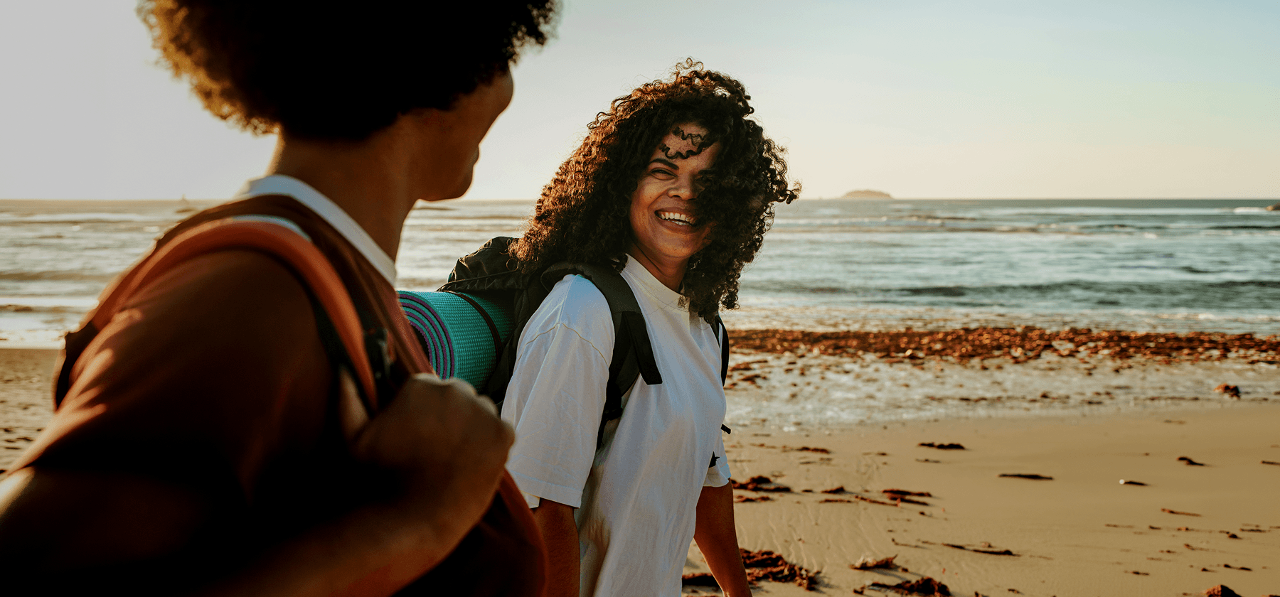 Two women with backpacks laughing together while walking along a sandy beach at sunset in Portugal.