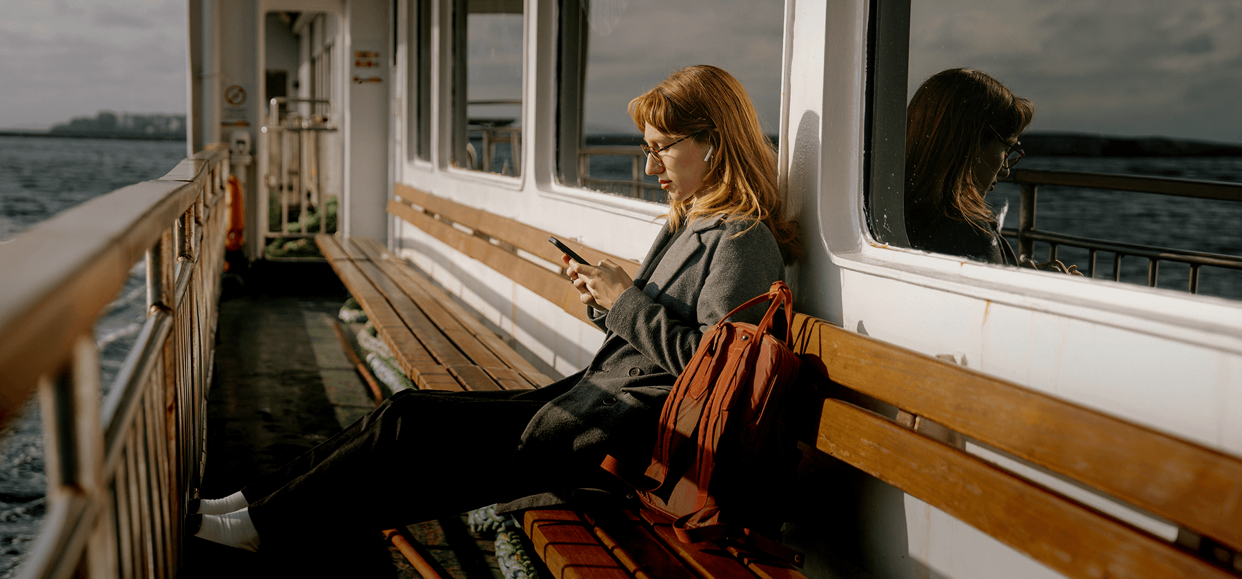A woman with red hair and glasses sitting on the outdoor deck of a ferry, looking at her smartphone with a red bag beside her in Sweden.