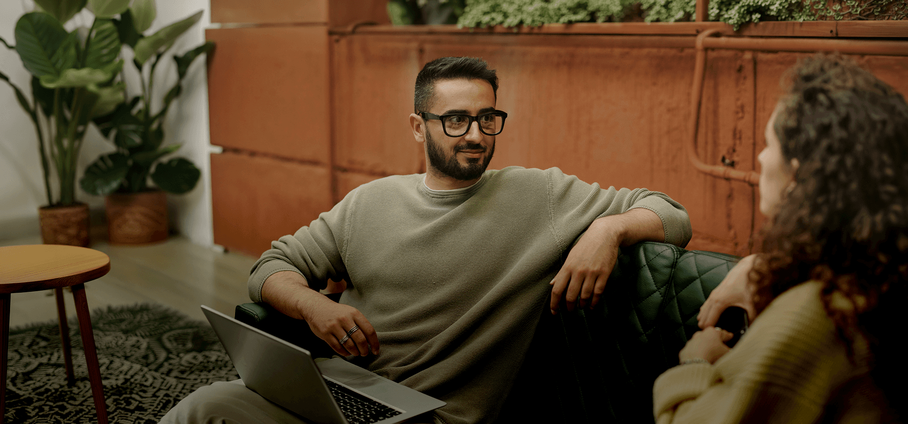 A bearded man wearing glasses sitting on a sofa with a laptop, engaged in conversation with someone across from him in France.