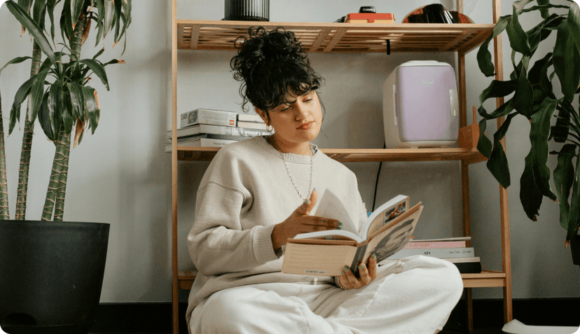 A girl sitting on a floor of her apartment next to a bookshelf, reading a book and relaxing.