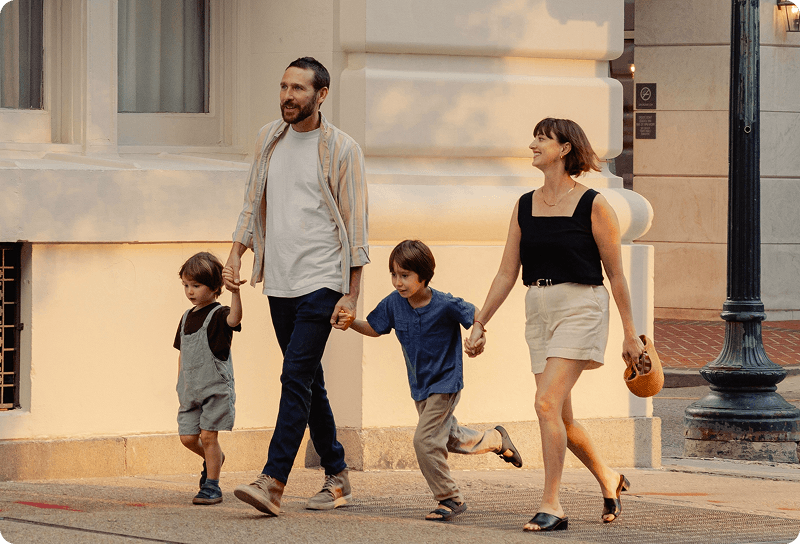 A family of four holding hands and walking together along a sunlit city street, with two young children between their parents in Germany.