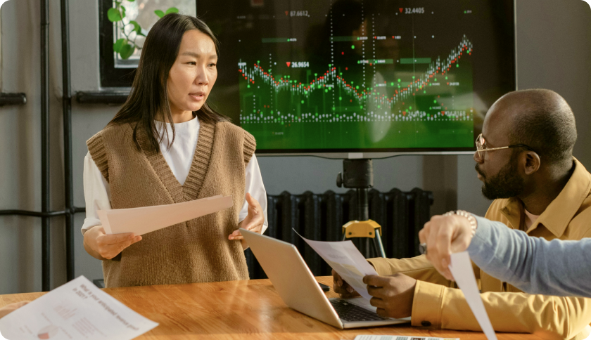 A consultant talking to her client during a business meeting. The tv screen behind her shows graphs.
