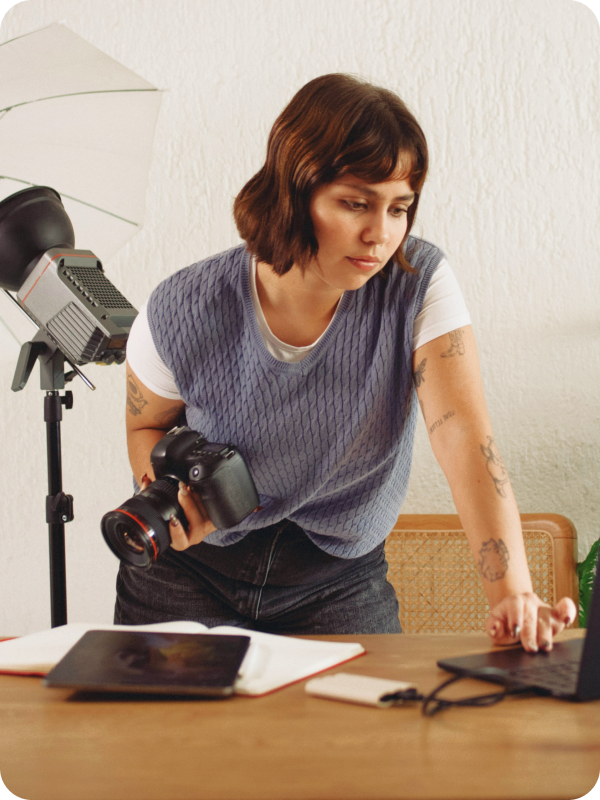 Photographer standing in front of her desk, holding a camera and checking her work on a laptop. Photoshoot equipment is seen in the background of the image.