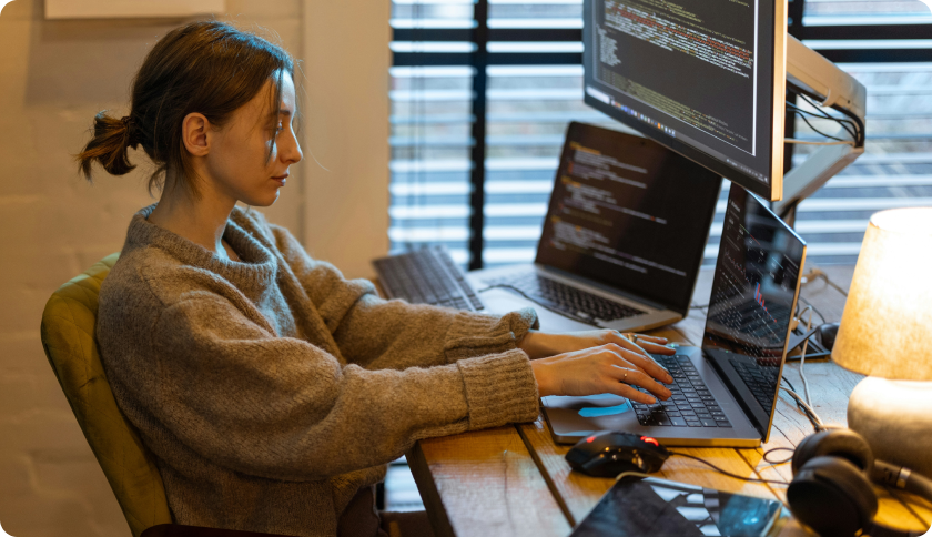 An IT freelancer working at her desk in front of 2 laptops and a screen showing lines of code.