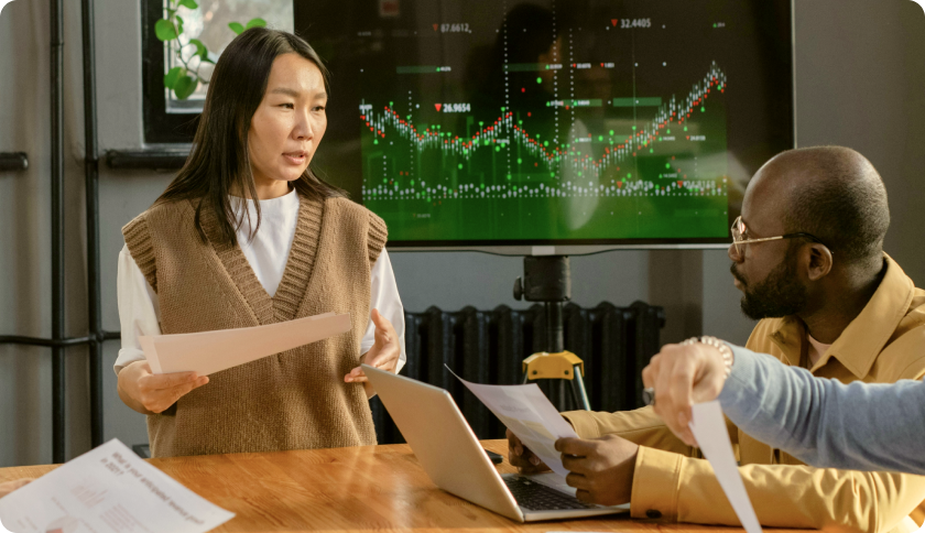 A consultant talking to her client during a business meeting. The tv screen behind her shows graphs.