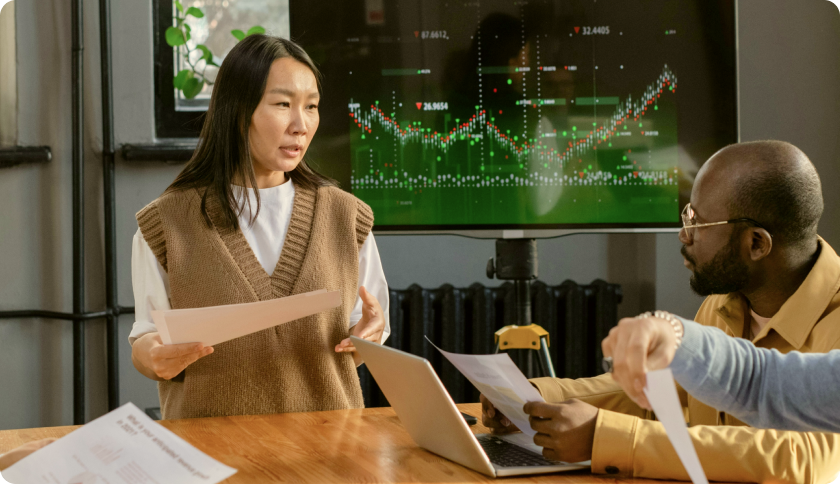 A consultant talking to her client during a business meeting. The tv screen behind her shows graphs.