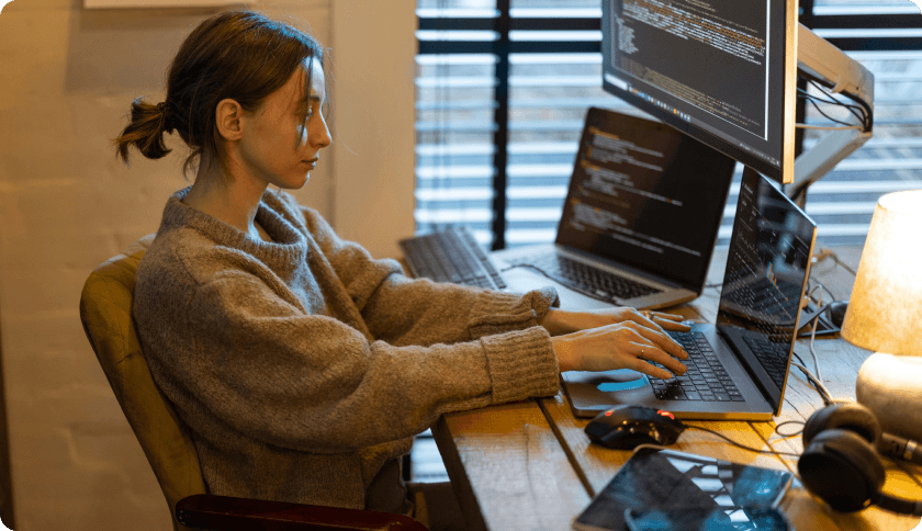 An IT freelancer working at her desk in front of 2 laptops and a screen showing lines of code.