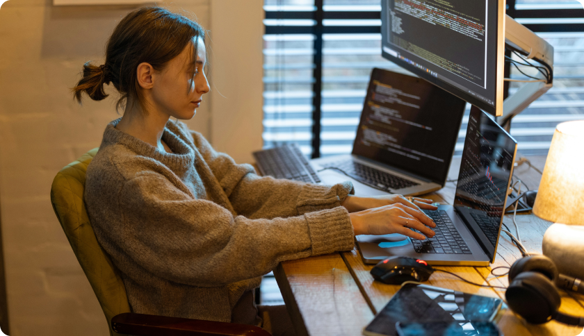 An IT freelancer working at her desk in front of 2 laptops and a screen showing lines of code.