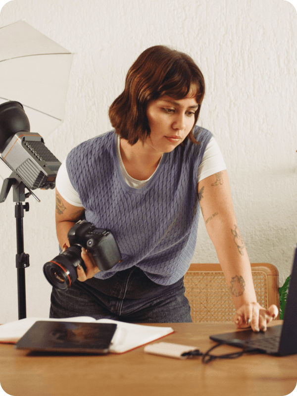 Photographer standing in front of her desk, holding a camera and checking her work on a laptop. Photoshoot equipment is seen in the background of the image.