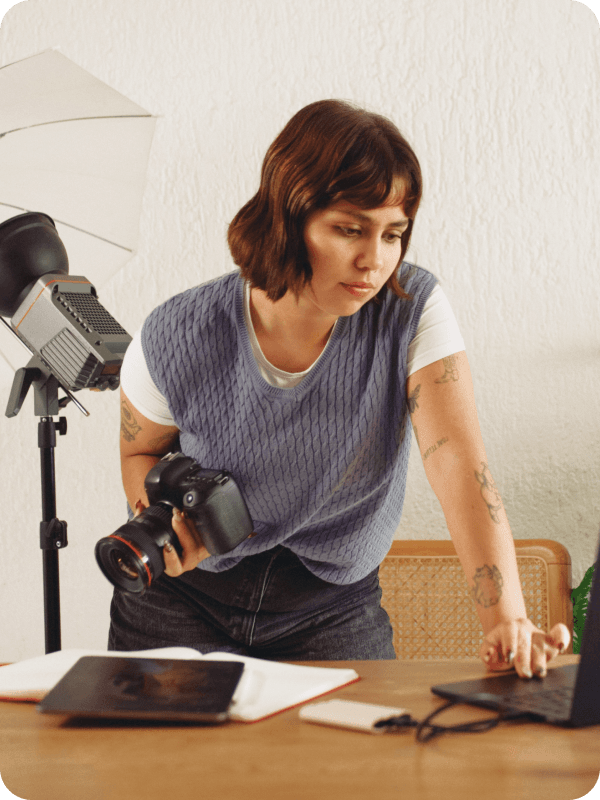 Photographer standing in front of her desk, holding a camera and checking her work on a laptop. Photoshoot equipment is seen in the background of the image.