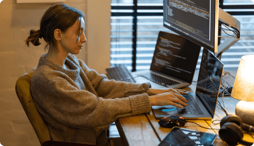 An IT freelancer working at her desk in front of 2 laptops and a screen showing lines of code.