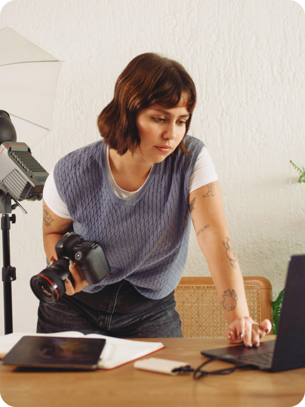 Photographer standing in front of her desk, holding a camera and checking her work on a laptop. Photoshoot equipment is seen in the background of the image.