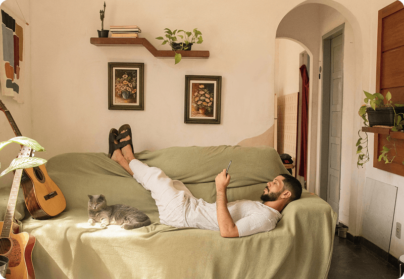 Person laying on a couch at their cosy home, looking at their phone with a cat laying by their side.