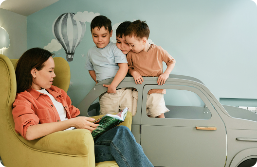 A woman sitting on an armchair reading a story to her 3 toddlers.